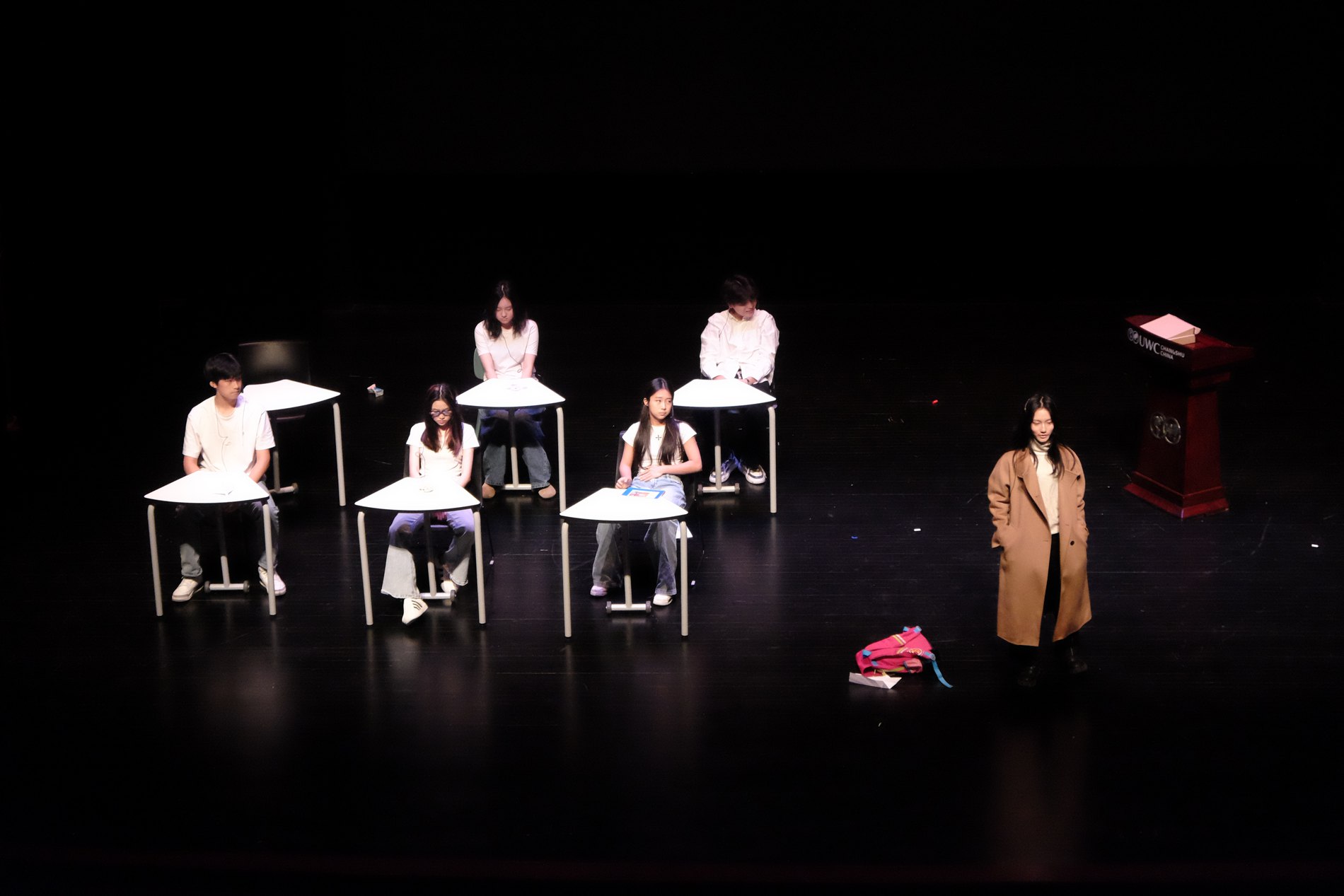 Ensemble seated at school desks in a dim stage lit scene from the original drama